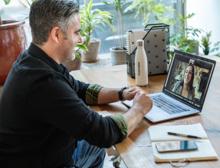 Man is setting at a table with his laptop open. On the screen is a woman. They seem to be in a meeting because he has a notebook, pen, water bottle, files, and his phone on the tabletop around him.